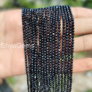 May include: Close-up of multiple strands of small, black, faceted beads. The beads are strung together, creating a dark, shimmering effect. The text "ShyaGems" is visible in the lower left corner.