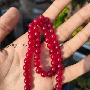 Könnte beinhalten: Eine Kette aus tiefroten, runden Edelsteinperlen. Die Perlen werden in einer Hand gehalten, wobei das Wort "Shyagems" im Hintergrund sichtbar ist. Die Perlen scheinen poliert zu sein und haben eine glatte Oberfläche. Das Gesamtthema ist Schmuck.