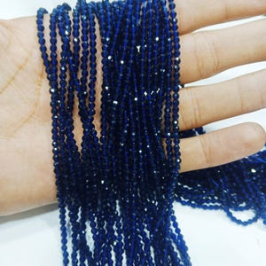 Può includere: Primo piano di fili di perline sfaccettate blu scuro. Le perline sono piccole e infilate insieme, creando un effetto scintillante. Le perline sono tenute in una mano, mostrando il loro colore e la loro consistenza. Ideale per la creazione di gioielli.