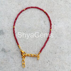 Op de afbeelding: Een delicate rode kralenarmband met goudkleurige accenten en een goudkleurige sluiting. De armband is geregen met kleine, ronde rode kralen en heeft de tekst "ShyaGems".