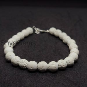 May include: White beaded bracelet with a textured surface. The bracelet is made of round beads and has a silver-coloured clasp. The text "Shya Gems" is visible on the left side of the image.
