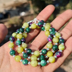 May include: A three-strand bracelet featuring round beads in shades of green, yellow, and purple. The beads are strung on a silver-colored chain. The bracelet is held in a hand.