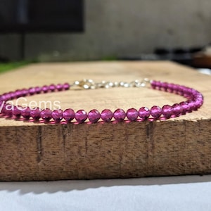 Può includere: Un delicato braccialetto con piccole perline sfaccettate color magenta infilate insieme. Il braccialetto ha una chiusura e una catena color argento. L'immagine è ambientata su una superficie di legno, con il testo "ShyaGems" visibile.