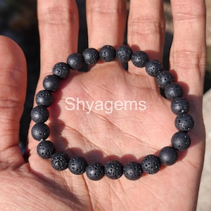 Puede incluir: Una pulsera de cuentas de piedra de lava negra. La pulsera está hecha de cuentas redondas y porosas. El texto "Shygems" está impreso en blanco en la imagen. La pulsera se muestra en la mano de una persona.