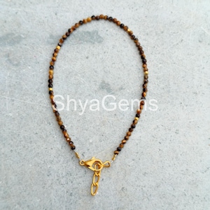 Può includere: Un braccialetto delicato con piccole perline rotonde di occhio di tigre marrone. Il braccialetto ha una chiusura e una catena color oro. Il testo "ShyaGems" è visibile nell'immagine.