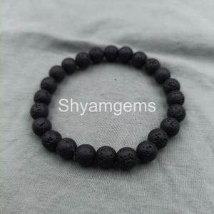 Può includere: Un braccialetto con perline di pietra lavica nera. Il braccialetto presenta perline rotonde e porose infilate insieme. Il testo "Shyamgems" è stampato in bianco al centro dell'immagine. Il braccialetto è esposto su una superficie grigio chiaro.