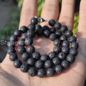 Puede incluir: Un collar de cuentas de piedra de lava negra. El collar está compuesto por cuentas redondas y porosas unidas por un cierre plateado. Las cuentas tienen una superficie rugosa y texturizada y son de color carbón oscuro.