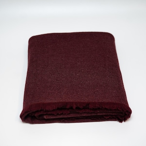 Puede incluir: Manta burdeos doblada con un borde con flecos. La manta tiene una textura tejida y está hecha de un material suave. La manta doblada está sobre un fondo blanco. Esta manta es adecuada para la decoración del hogar.
