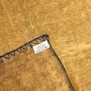 以下が含まれることがあります： 黄色の織物生地のクローズアップ。青いラベルには「Yak Wool Handmade in Nepal Machine Washable」と記載されています。ラベルには、洗濯、乾燥、アイロンがけのシンボルが記されたお手入れ方法が記載されています。