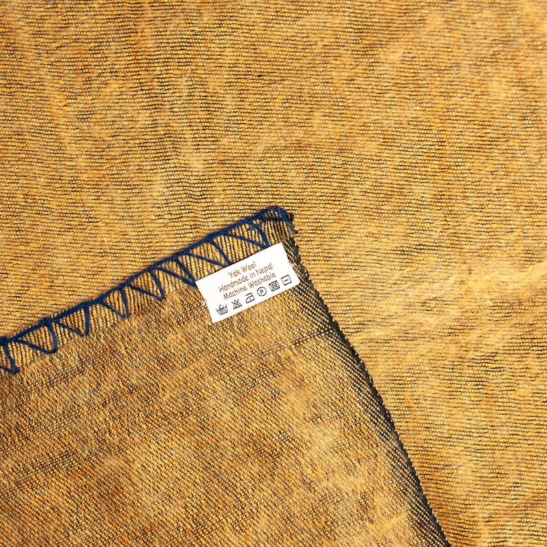 以下が含まれることがあります： 黄色の織物生地のクローズアップ。青いラベルには「Yak Wool Handmade in Nepal Machine Washable」と記載されています。ラベルには、洗濯、乾燥、アイロンがけのシンボルが記されたお手入れ方法が記載されています。