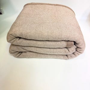 K&ouml;nnte beinhalten: Gefaltete beige Decke mit Fischgr&auml;tenmuster. Die Decke hat einen dunkelbraunen Rand und ist ordentlich gestapelt. Die weichen, neutralen T&ouml;ne deuten auf ein gem&uuml;tliches Heimtextil hin.