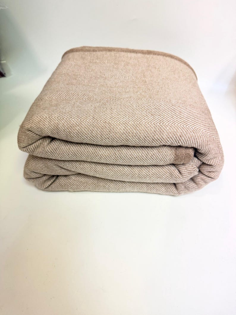 K&ouml;nnte beinhalten: Gefaltete beige Decke mit Fischgr&auml;tenmuster. Die Decke hat einen dunkelbraunen Rand und ist ordentlich gestapelt. Die weichen, neutralen T&ouml;ne deuten auf ein gem&uuml;tliches Heimtextil hin.