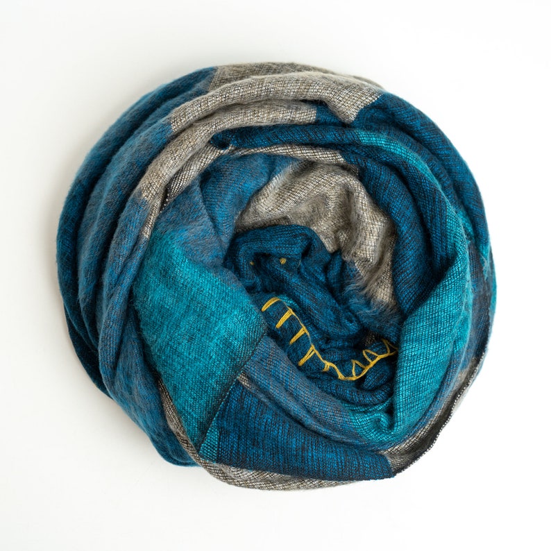 May include: A blue and gray plaid scarf is rolled up into a circle. The scarf is made of a soft, fuzzy material.