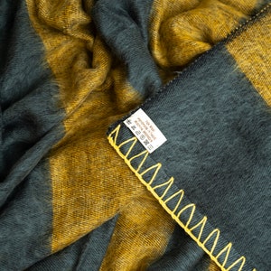 May include: A soft, striped throw blanket in shades of mustard yellow and dark grey. The blanket has a yellow stitched border and a care label that reads "100% Wool, Made in India, Wool Wash Only".