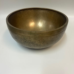 May include: A bronze-coloured singing bowl with a rounded shape. The bowl has a textured surface and a dark rim. The interior reflects light, and the bowl appears to be made of metal. The bowl is likely used for meditation or sound healing.