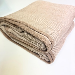 K&ouml;nnte beinhalten: Gefaltete beige Decke mit Fischgr&auml;tenmuster. Die Decke hat einen dunkleren braunen Rand und ist ordentlich gestapelt. Die neutrale Farbpalette und Textur deuten auf ein gem&uuml;tliches Heimtextil hin.