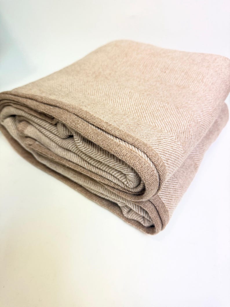 K&ouml;nnte beinhalten: Gefaltete beige Decke mit Fischgr&auml;tenmuster. Die Decke hat einen dunkleren braunen Rand und ist ordentlich gestapelt. Die neutrale Farbpalette und Textur deuten auf ein gem&uuml;tliches Heimtextil hin.