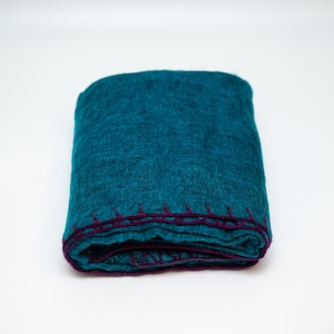 May include: Folded teal blanket with a dark purple stitched border. The blanket is made of a textured fabric and is neatly folded. The color is a vibrant teal, and the stitching provides a contrasting accent.