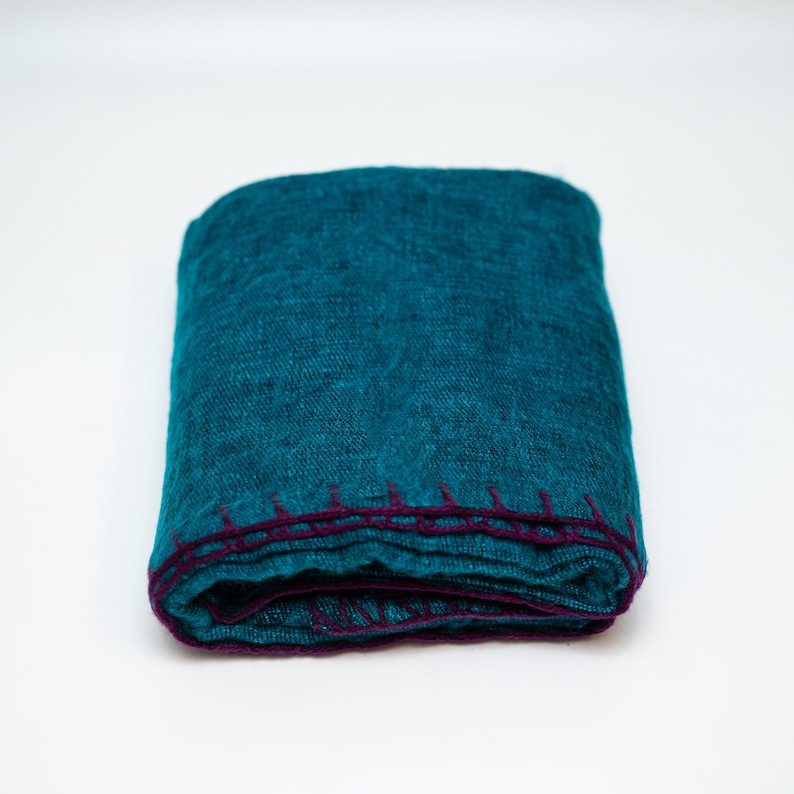 May include: Folded teal blanket with a dark purple stitched border. The blanket is made of a textured fabric and is neatly folded. The color is a vibrant teal, and the stitching provides a contrasting accent.