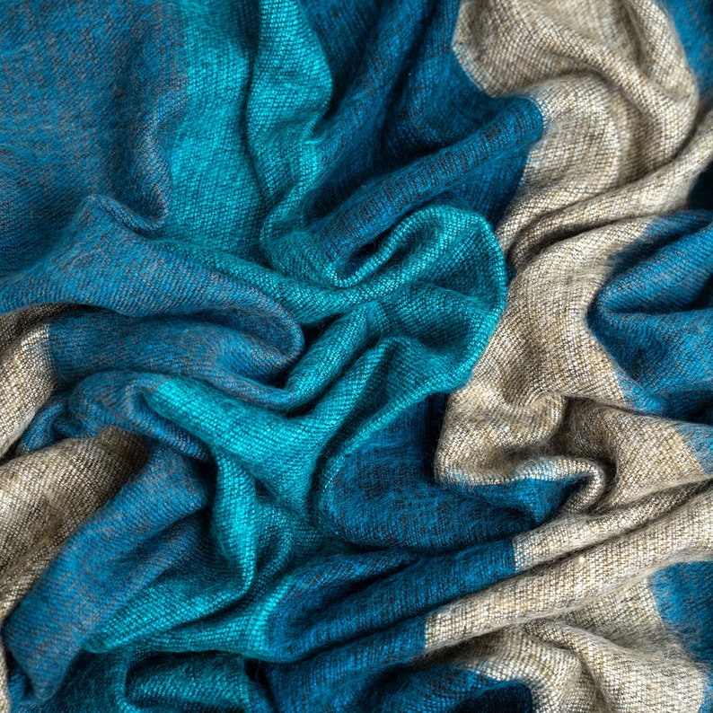May include: A soft, blue and beige striped scarf with a textured weave. The scarf is folded and draped in a way that shows the different colors and textures.