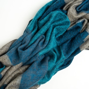 May include: A soft, blue and gray striped scarf made of wool. The scarf is folded and draped on a white surface.