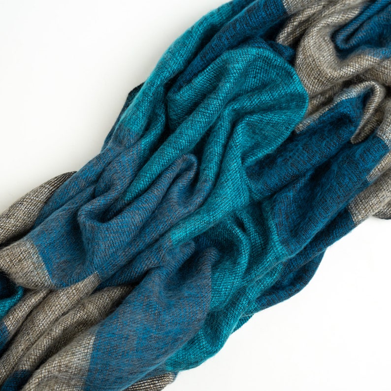 May include: A soft, blue and gray striped scarf made of wool. The scarf is folded and draped on a white surface.