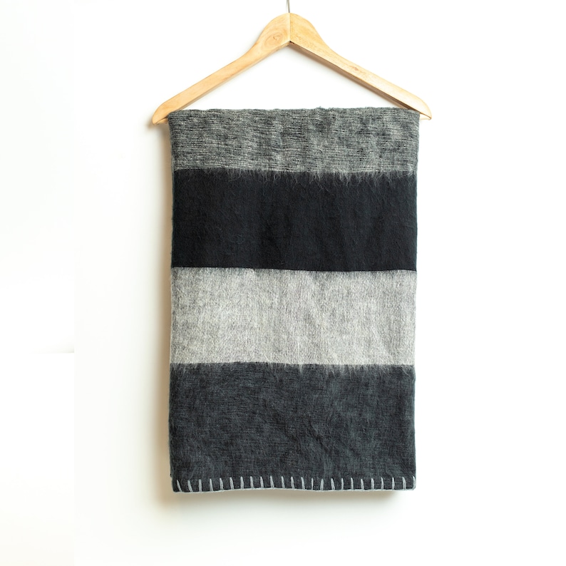 May include: A black and gray striped throw blanket hanging on a wooden hanger. The blanket has a fuzzy texture and is made of wool.
