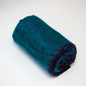 May include: Rolled teal-colored blanket with a textured weave. The blanket has a deep purple stitched border. The blanket is rolled up, showing the texture and color variations. The blanket is on a white background.