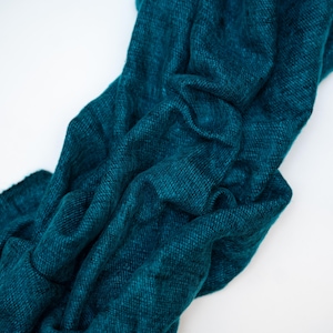 May include: A teal-colored scarf made of a soft, woven material. The scarf is draped and folded, showing the texture and depth of the fabric. The background is plain white, which highlights the scarf's color and texture.