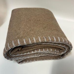Puede incluir: Manta marrón doblada con costuras blancas en los bordes. La manta está hecha de una tela texturizada y parece cálida y acogedora. Las costuras añaden un elemento decorativo.