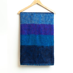 May include: A blue and purple striped wool blanket hanging on a wooden hanger. The blanket has a fuzzy texture and is fringed on the bottom.