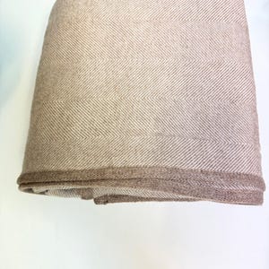 K&ouml;nnte beinhalten: Gefaltete beige Decke mit Fischgr&auml;tenmuster. Die Decke hat einen dunkleren braunen Rand. Der Stoff scheint ein weiches, gewebtes Material zu sein, das f&uuml;r W&auml;rme und Komfort geeignet ist.