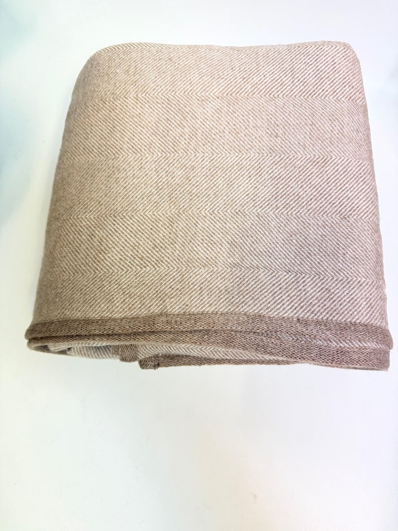 K&ouml;nnte beinhalten: Gefaltete beige Decke mit Fischgr&auml;tenmuster. Die Decke hat einen dunkleren braunen Rand. Der Stoff scheint ein weiches, gewebtes Material zu sein, das f&uuml;r W&auml;rme und Komfort geeignet ist.