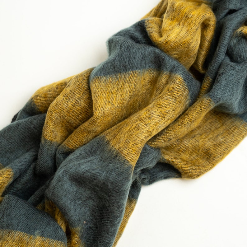 May include: A soft, striped throw blanket in shades of mustard yellow and dark teal. The blanket is made of a fuzzy, textured material and is folded in a loose, casual way.