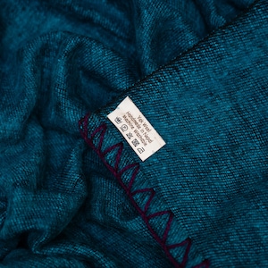 May include: A teal-colored textile with a textured appearance. The edge is finished with a deep purple, zig-zag stitch. A white tag with black text is visible, indicating the material and care instructions.