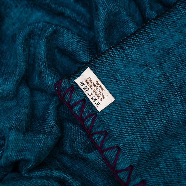 May include: A teal-colored textile with a textured appearance. The edge is finished with a deep purple, zig-zag stitch. A white tag with black text is visible, indicating the material and care instructions.