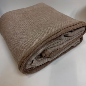 K&ouml;nnte beinhalten: Gefaltete braune Decke mit Fischgr&auml;tenmuster. Die Decke hat einen dunkleren braunen Rand und besteht aus einem weichen, gewebten Material. Die Decke ist ordentlich gefaltet und zeigt ihre Textur und ihr Design.