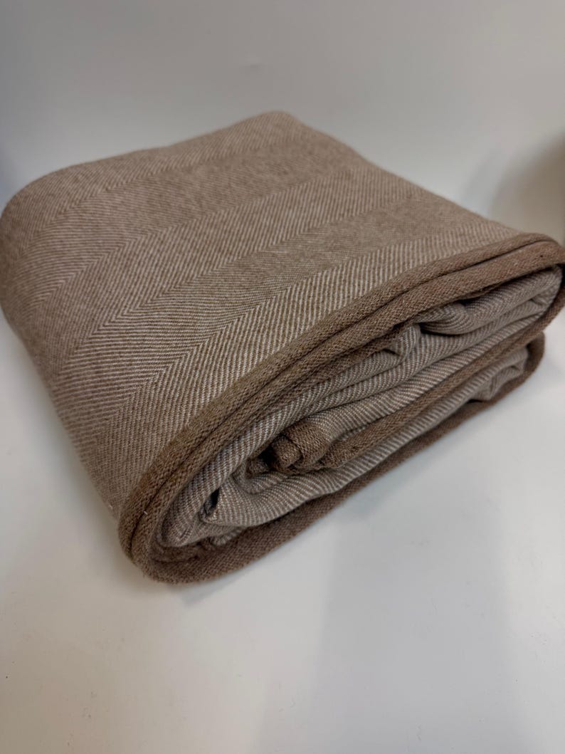 K&ouml;nnte beinhalten: Gefaltete braune Decke mit Fischgr&auml;tenmuster. Die Decke hat einen dunkleren braunen Rand und besteht aus einem weichen, gewebten Material. Die Decke ist ordentlich gefaltet und zeigt ihre Textur und ihr Design.