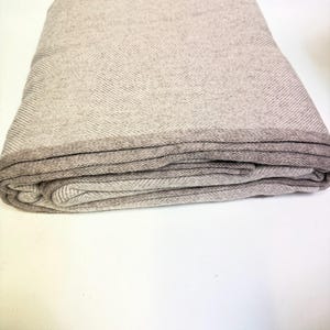 Op de afbeelding: Gevouwen beige deken met een visgraatpatroon. De deken heeft een donkerdere rand en is netjes opgevouwen, wat de textuur en het ontwerp laat zien. Het neutrale kleurenpalet suggereert een gezellig en veelzijdig huistextiel.