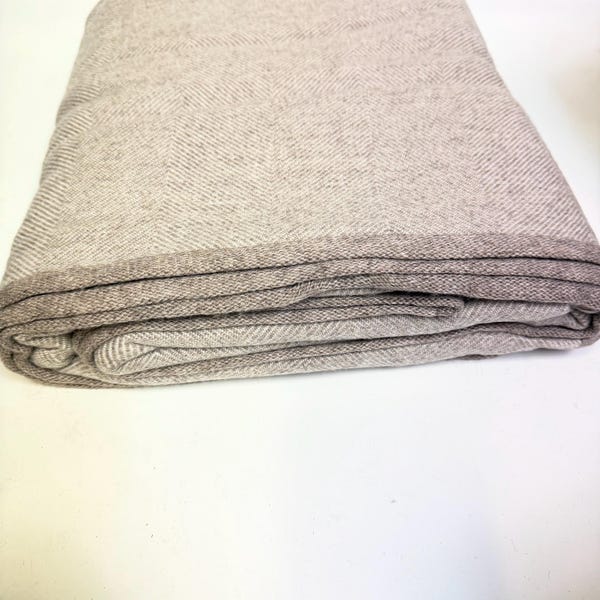 Può includere: Coperta beige piegata con motivo a spina di pesce. La coperta ha un bordo più scuro ed è ben piegata, mettendo in mostra la trama e il design. La palette di colori neutri suggerisce un tessuto per la casa accogliente e versatile.