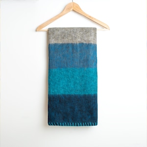 May include: A striped throw blanket with a gray, blue, and teal color scheme. The blanket is hanging on a wooden hanger against a white background.