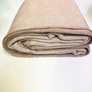 K&ouml;nnte beinhalten: Gefaltete beige Decke mit Fischgr&auml;tenmuster. Die Decke hat einen dunkleren beigen Rand und ist ordentlich gestapelt. Der Stoff wirkt weich und warm, geeignet f&uuml;r ein Bett oder Sofa.