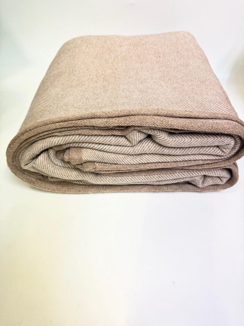 K&ouml;nnte beinhalten: Gefaltete beige Decke mit Fischgr&auml;tenmuster. Die Decke hat einen dunkleren beigen Rand und ist ordentlich gestapelt. Der Stoff wirkt weich und warm, geeignet f&uuml;r ein Bett oder Sofa.