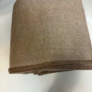 K&ouml;nnte beinhalten: Gefaltete braune und beige Decke mit Fischgr&auml;tenmuster. Die Decke hat ein klassisches Fischgr&auml;tenmuster in einer warmen, neutralen Farbgebung. Die Kanten sind sauber verarbeitet, was auf Qualit&auml;t hindeutet. Ideal f&uuml;r Heimdekoration und Komfort.