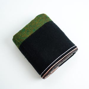 May include: A folded rectangular textile featuring a large solid black section, a contrasting green textured band with subtle brown flecks, and a thin decorative border with white and peach-coloured stripes along the bottom edge.