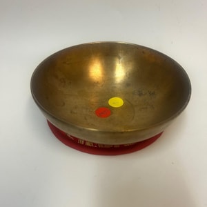 May include: A brass singing bowl with a red cushion. The bowl has a worn, aged look and is resting on a red cushion with gold embroidery.