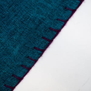 May include: Close-up of a teal fabric with a textured appearance. The edge is finished with a deep purple stitching. The fabric is set against a white background, highlighting the color and texture of the material.