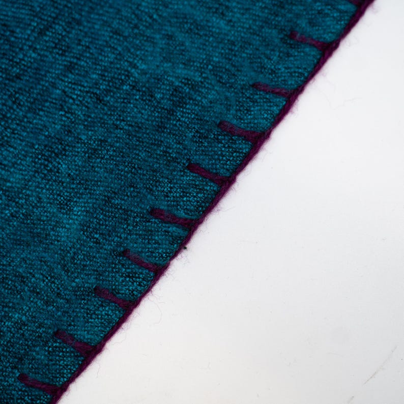 May include: Close-up of a teal fabric with a textured appearance. The edge is finished with a deep purple stitching. The fabric is set against a white background, highlighting the color and texture of the material.