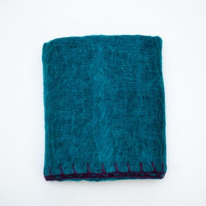 May include: A folded teal blanket with a textured appearance. The edges are finished with a dark purple stitched border. The blanket is made of a soft, possibly woven material, and is displayed against a white background.