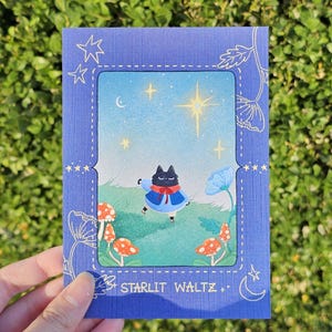 May include: A blue greeting card with a whimsical illustration of a black cat in a blue coat dancing in a field with mushrooms and flowers. The card has the text "Starlit Waltz".
